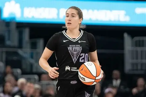 A WNBA player in a Golden State Valkyries uniform dribbling the basketball during a pre-season game, with arena crowd visible in the background.