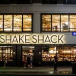 Shake Shack shares drop — Shake Shack restaurant exterior with illuminated signage at night, showing the premium burger chain's branding clearly visible to passing customers.
