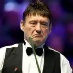Snooker legend Jimmy White holding a cue at a snooker tournament, smiling at the camera.